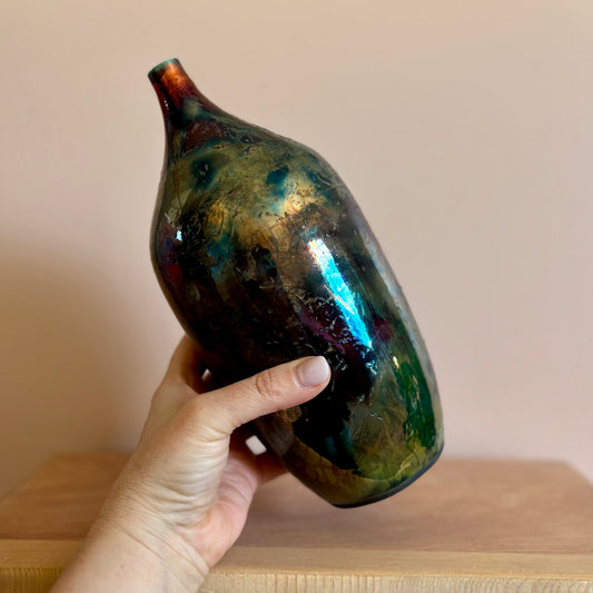 a hand holding a porcelain raku-fired vase in iridescent hues of green and purple. Made by Hillit Wahlberg