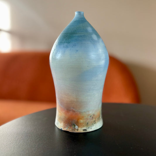 A saggar fired porcelain vase with a round guitar shaped body and a tiny neck in light shades of blue and brown. Made by ceramic artist Hillit Wahlberg