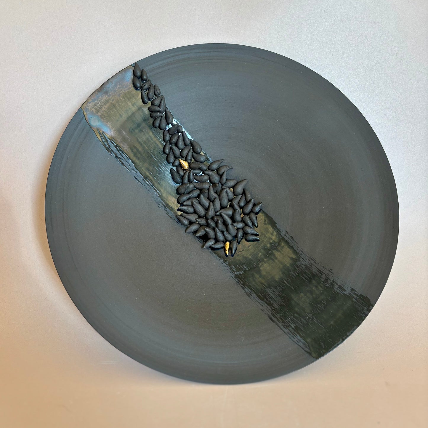 A circular sculpture or decorative bowl in dark gray and black, featuring a raised diagonal band of textured black seed-like forms on a beige wall background.