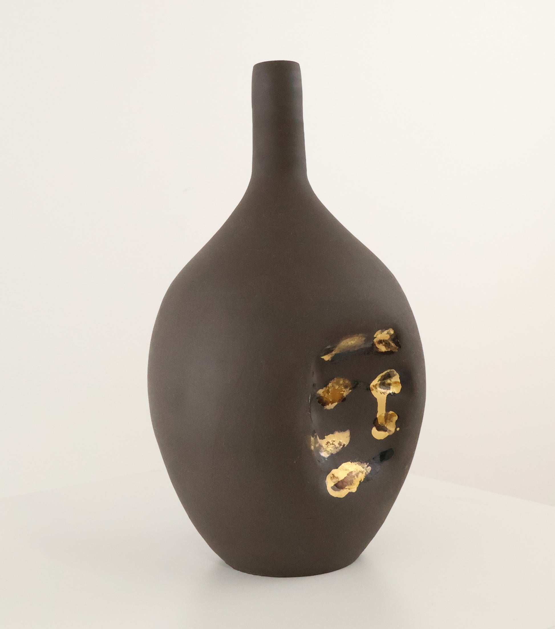 A dark brown matte ceramic vase with a short neck and irregular organic silhouette, featuring a recessed area on the side filled with textured 22k gold leaf, set against a plain off-white background.