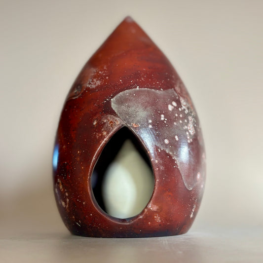 A teardrop-shaped ceramic sculpture with a glossy dark reddish-brown exterior, white speckled glaze details, and a central cutout revealing a pale cream interior, set against a neutral beige background.
