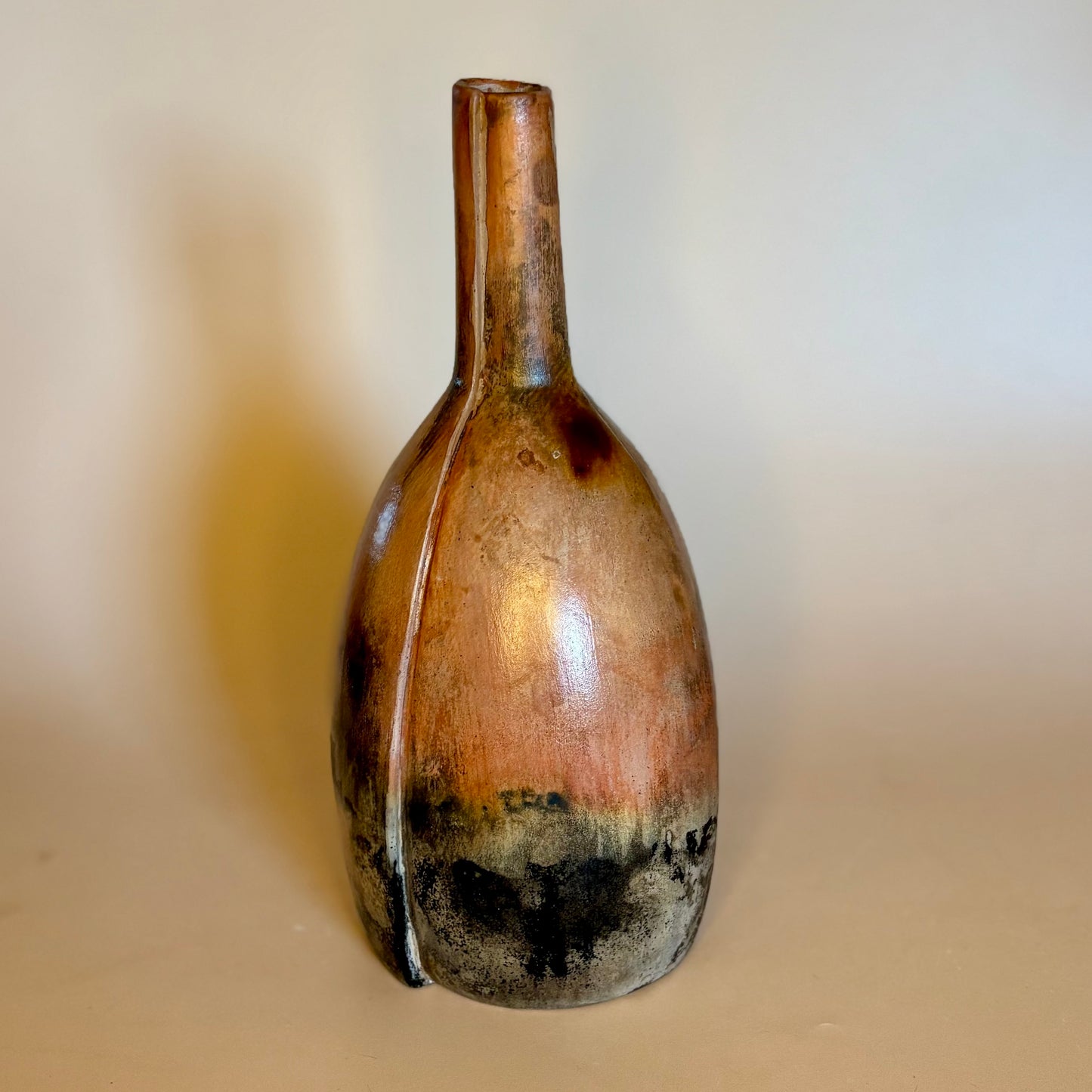 A rustic ceramic vase with a narrow neck and bulbous body, featuring a glossy finish with brown, coppery, and black glaze variations, set against a plain beige background.