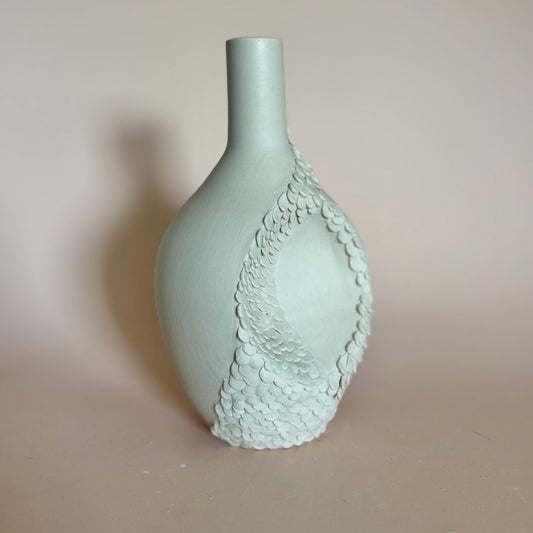 A pale beige ceramic vase with a textured, scale-like pattern on one side, set against a plain beige background.