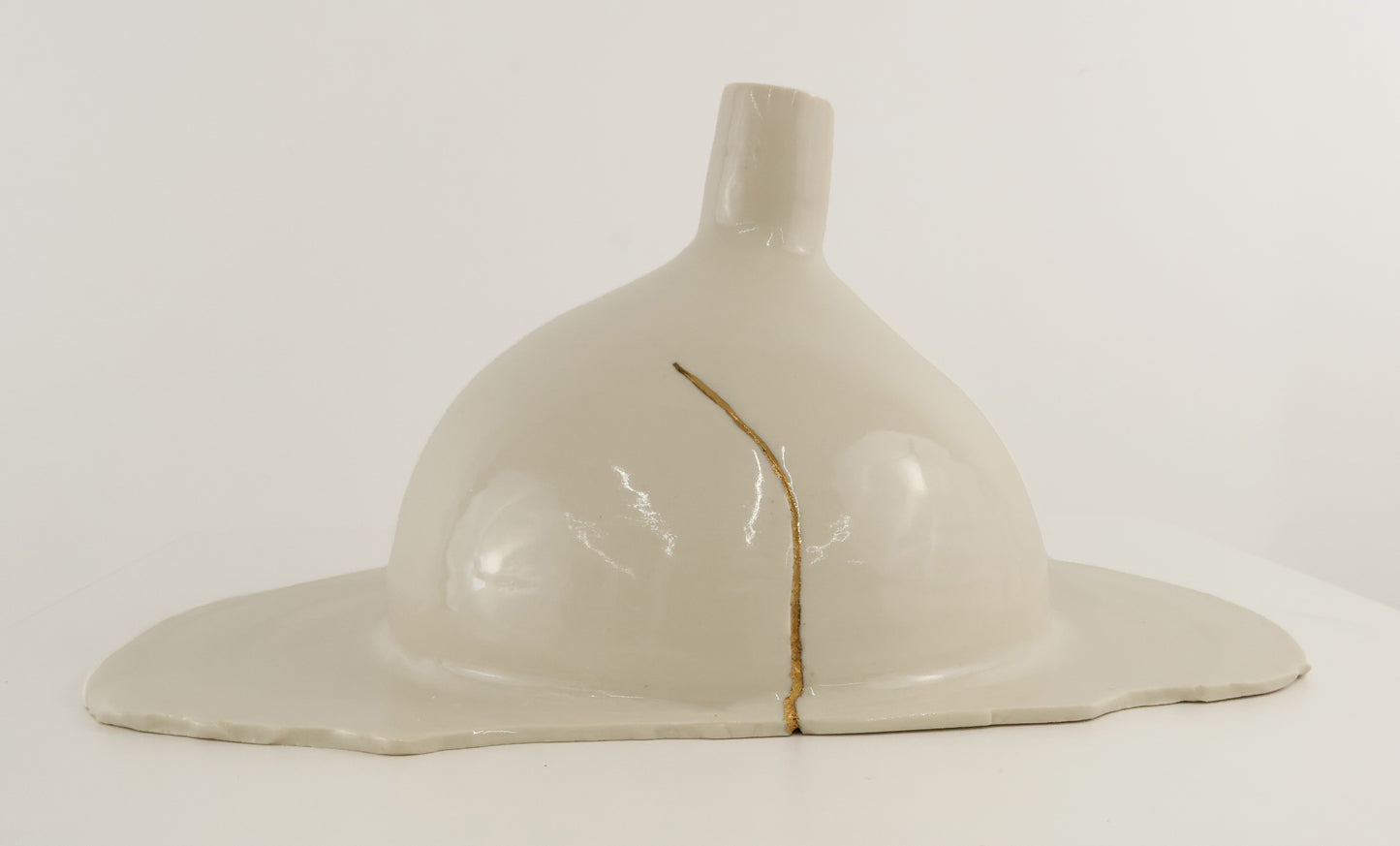 A ceramic sculpture of a stylized dome or helmet-like form with a small neck and wide flared base, shown on a plain white studio background. The piece is glossy off-white/cream with a thin vertical gold line running down the center.