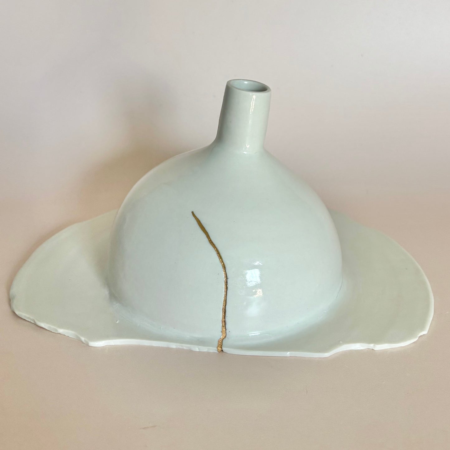 A handmade ceramic vase with a wide, shallow saucer-like base and a narrow neck, glazed in a pale celadon green with a single vertical gold fiber reinforcement crack running down the center, set against a solid light beige background.