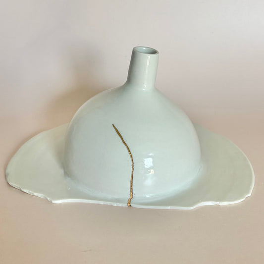 A handmade ceramic vase with a wide, shallow saucer-like base and a narrow neck, glazed in a pale celadon green with a single vertical gold fiber reinforcement crack running down the center, set against a solid light beige background.
