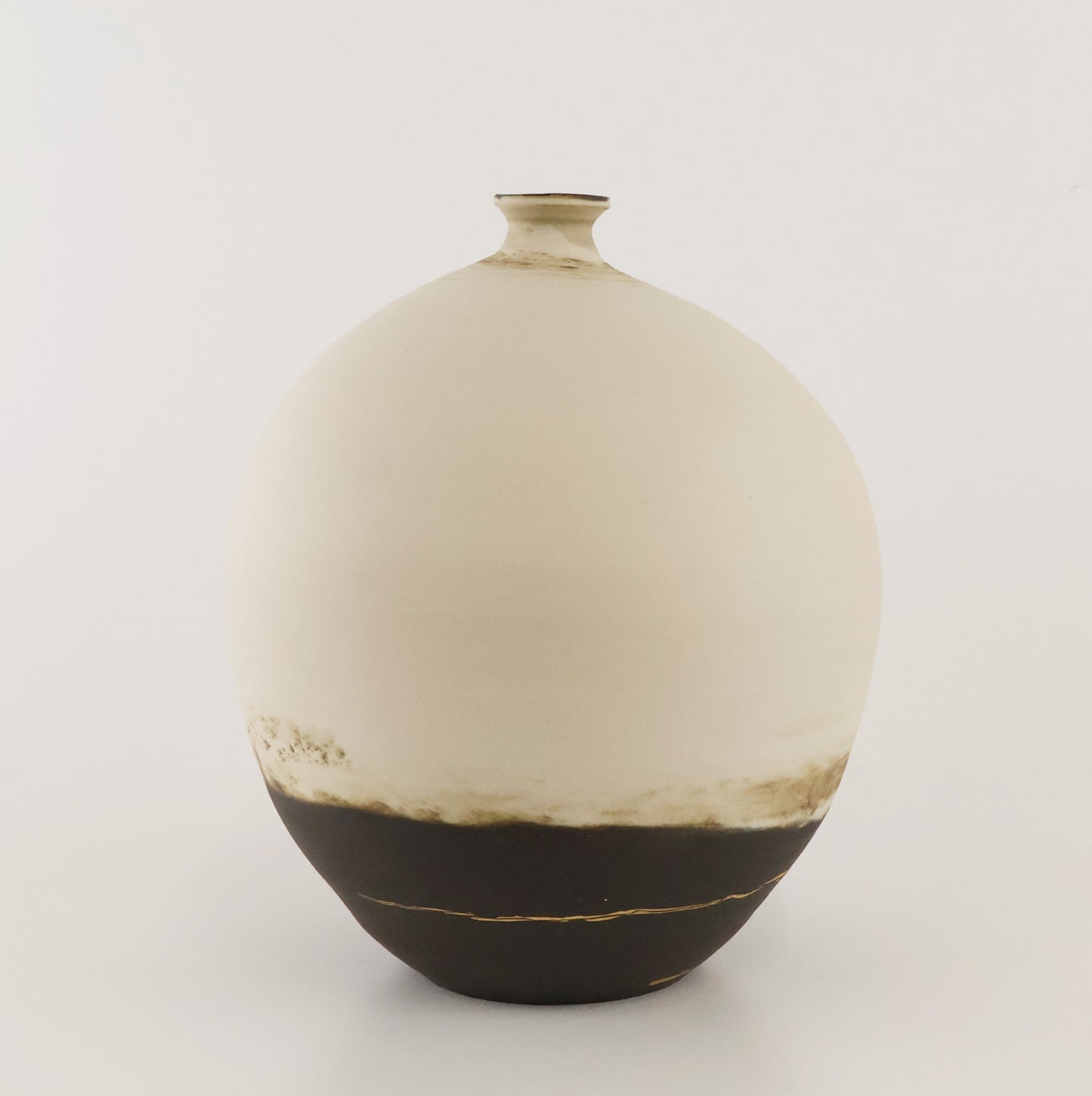 A ceramic vase with a rounded, bulbous shape, featuring a matte off-white upper body that fades into a dark brown base with subtle 22k gold glaze accents, set against a plain light gray background.