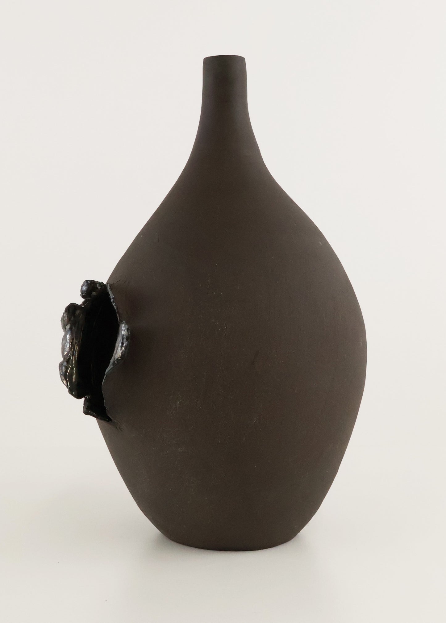 A matte dark brown ceramic vase with a narrow neck and a protruding sculptural black element on the side, photographed against a plain light background.