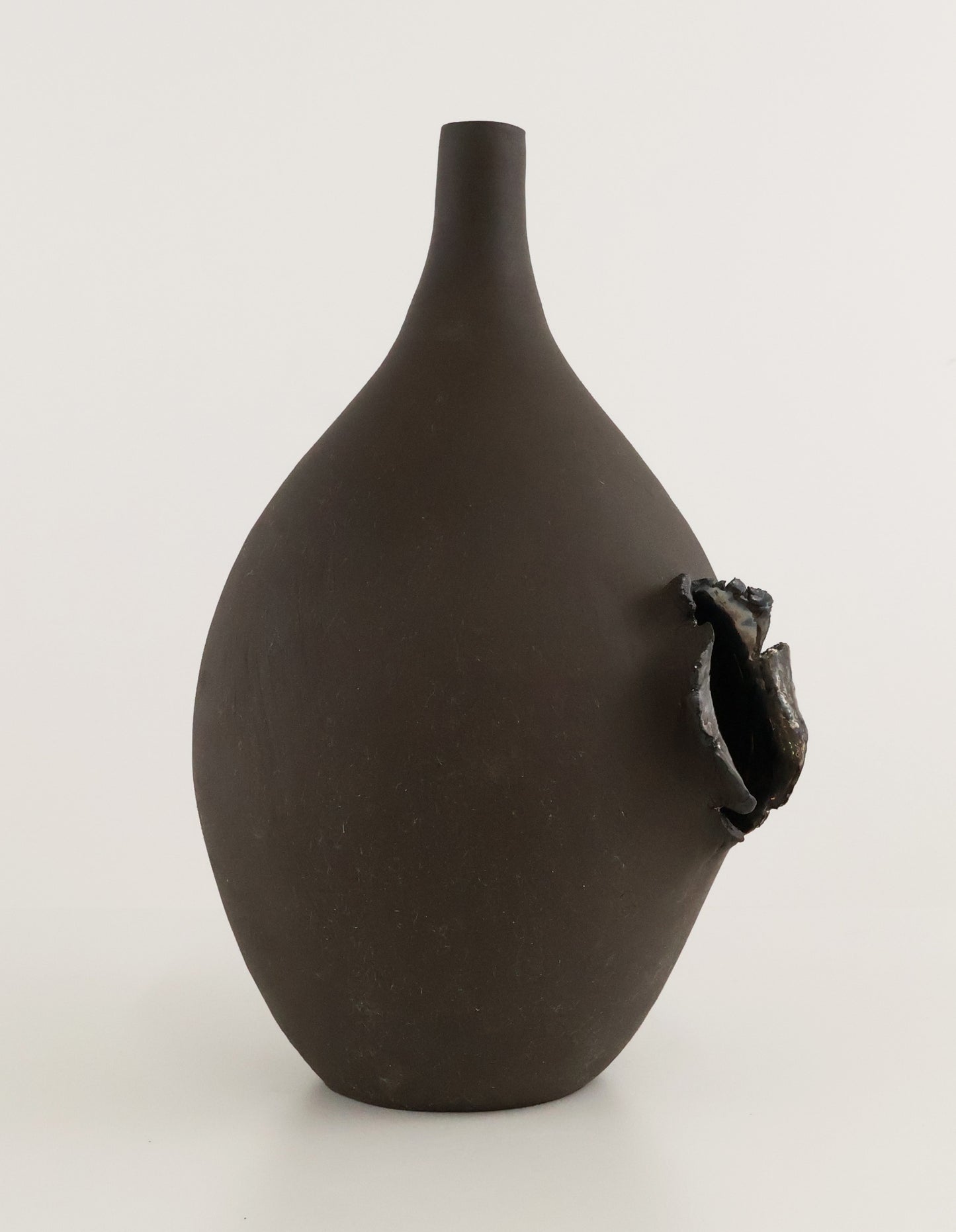 A dark brown matte ceramic vase with a narrow neck and a textured, organic protruding opening on the side, set against a plain light gray background.