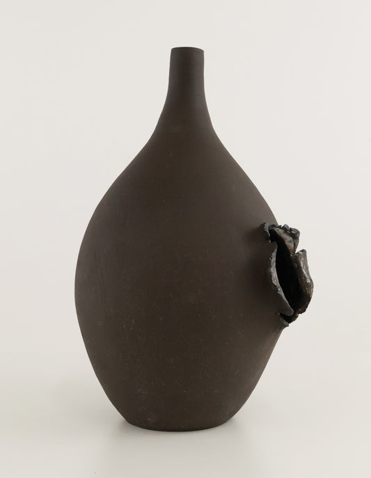 A dark brown matte ceramic vase with a narrow neck and a textured, organic protruding opening on the side, set against a plain light gray background.
